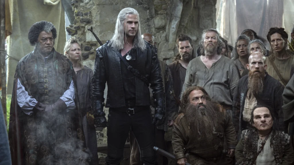 Eine Gruppe von Menschen in mittelalterlichen Gewändern steht in einer steinernen Umgebung. Im Zentrum steht eine Person mit langen, weissen Haaren in schwarzer Lederkleidung. Umgeben von verschiedenen Charakteren, einige blicken in die Ferne, andere direkt in die Kamera.