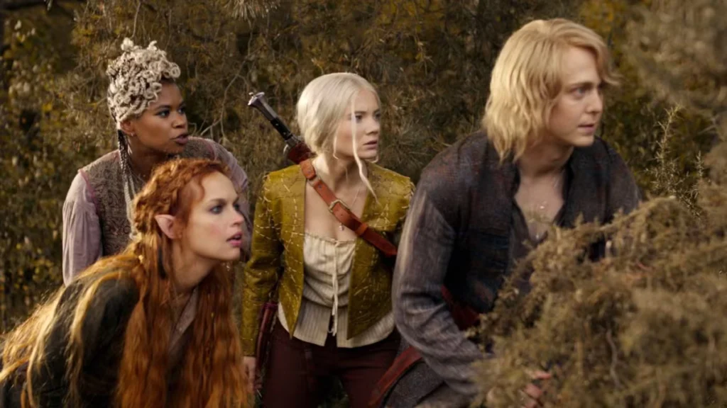 Vier Personen in mittelalterlichen Fantasiekostümen stehen aufmerksam hinter Büschen. Links vorne eine Person mit spitzen Ohren und langen, rotbraunen Haaren. In der Mitte tragen zwei Personen blonde bzw. graue Haare und Umhänge. Der Hintergrund zeigt einen bewaldeten Bereich.