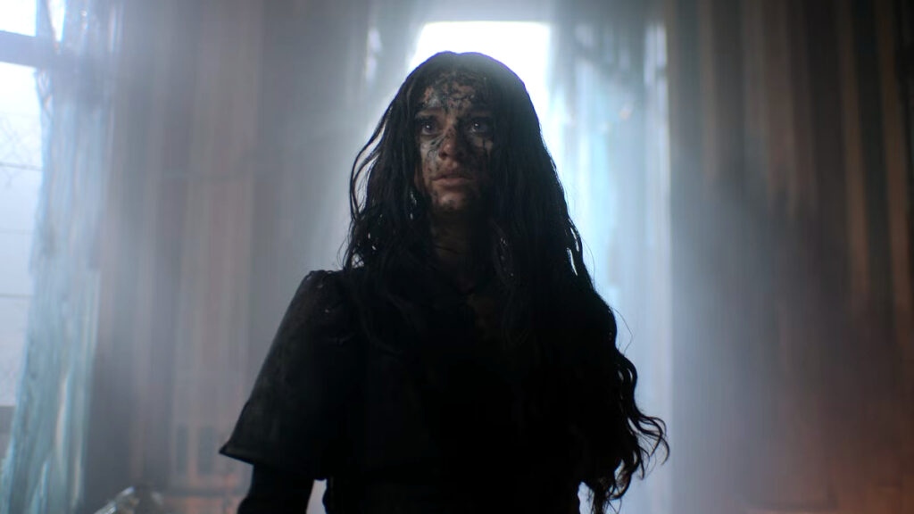 Eine Person mit schmutzigem Gesicht und langen, zerzausten Haaren steht in einem düsteren, vernebelten Raum. Das Licht scheint von hinten und hebt die Silhouette hervor. Die Atmosphäre ist geheimnisvoll und dramatisch, passend zu einer TV-Streamingserie mit einem dunklen Thema.