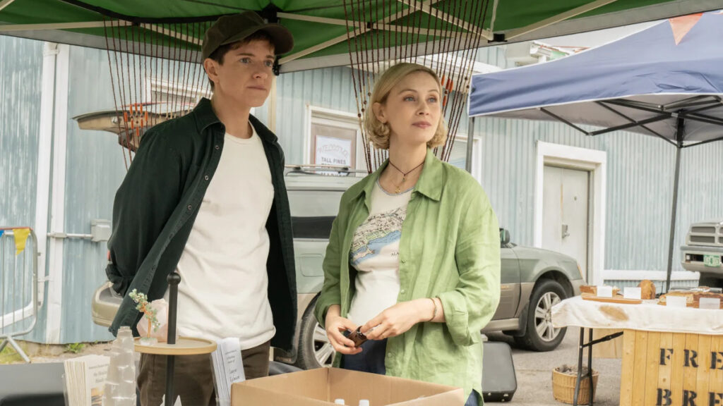 Zwei Personen stehen unter einem grünen Pavillon auf einem Markt. Sie tragen lässige Kleidung; eine Person trägt eine Mütze. Im Vordergrund steht ein Tisch mit Büchern und Verkaufsartikeln, im Hintergrund ist ein Auto sichtbar.