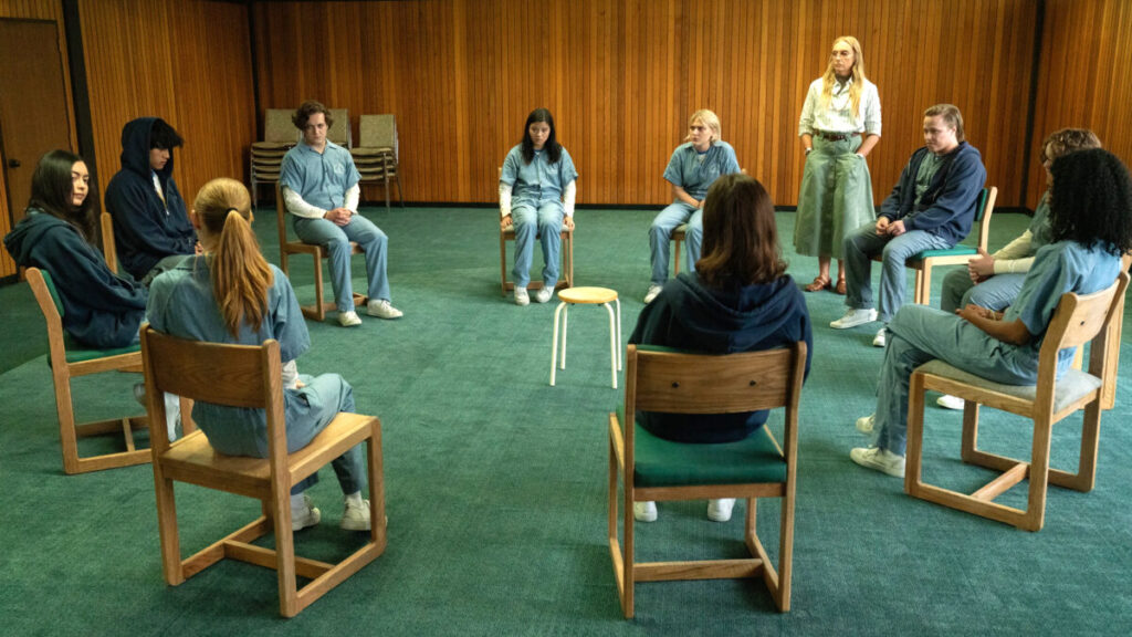 Eine Gruppe von jungen Menschen sitzt in einem Stuhlkreis in einem Raum mit Holzverkleidung. Einige tragen einheitliche Kleidung. Eine Person steht, während die anderen sitzen und zuhören. Ein leerer Stuhl steht in der Mitte des Kreises.