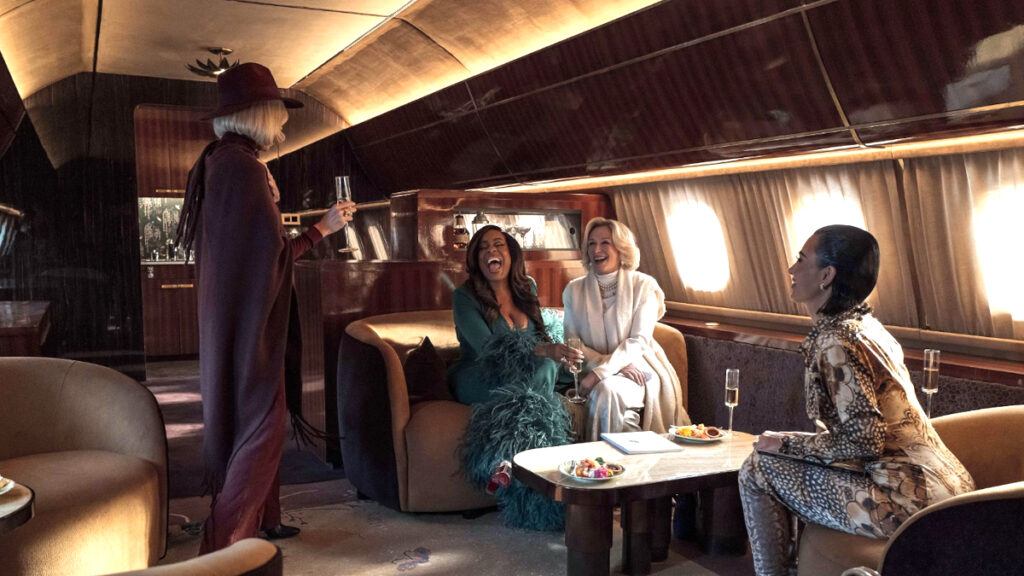 Vier elegant gekleidete Frauen sitzen in einem luxuriösen Privatjet. Eine steht und hält ein Getränk, während die anderen auf Sofas um einen Tisch lachen, auf dem Snacks liegen. Das Ambiente ist luxuriös und stilvoll, mit gedämpfter Beleuchtung und Holzverkleidung im Hintergrund.