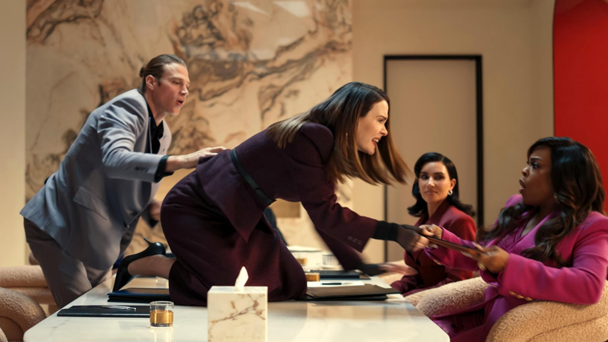 Vier Personen in einem modernen Büro. Eine Frau im lila Anzug kniet auf dem Tisch und streitet mit einer sitzenden Frau in rosa Kleidung. Zwei weitere Personen beobachten die Szene. Im Hintergrund eine kunstvolle Wanddekoration. Spannungsgeladene Atmosphäre.