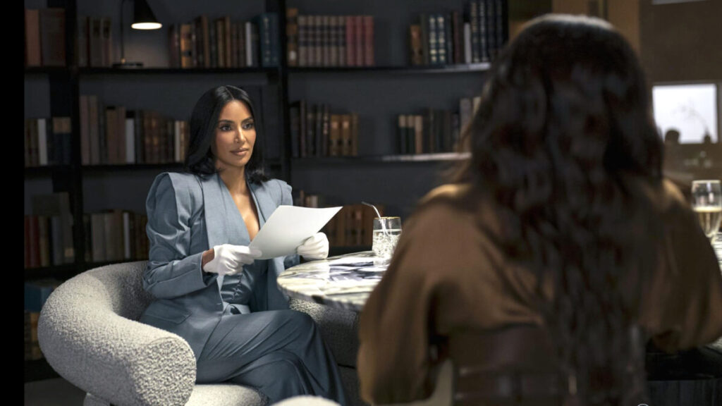 Eine Frau in einem schicken Anzug sitzt an einem Tisch in einer stilvollen Bibliothek. Sie hält ein Blatt Papier in behandschuhten Händen und blickt auf eine andere Person, die ihr gegenüber sitzt. Im Hintergrund sind Bücherregale sichtbar.