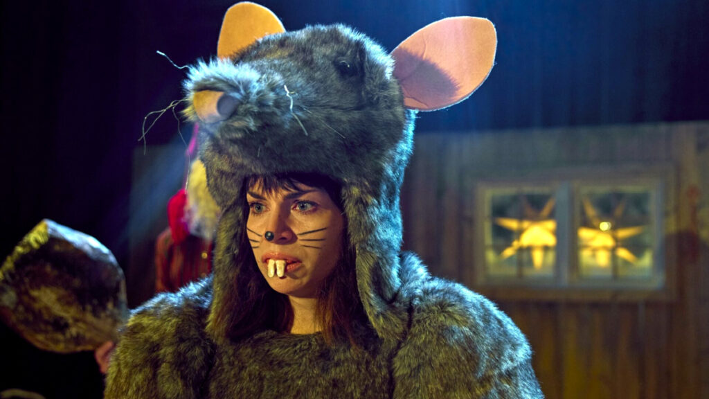 Eine Person in einem realistischen Rattenkostüm mit grossen Ohren und Schnurrhaaren. Sie trägt Gesichtsbemalung mit Rattenzähnen. Hinter ihr ist ein Fenster mit gelbem Licht zu sehen. Die Szene wirkt theatralisch und ist warm beleuchtet.