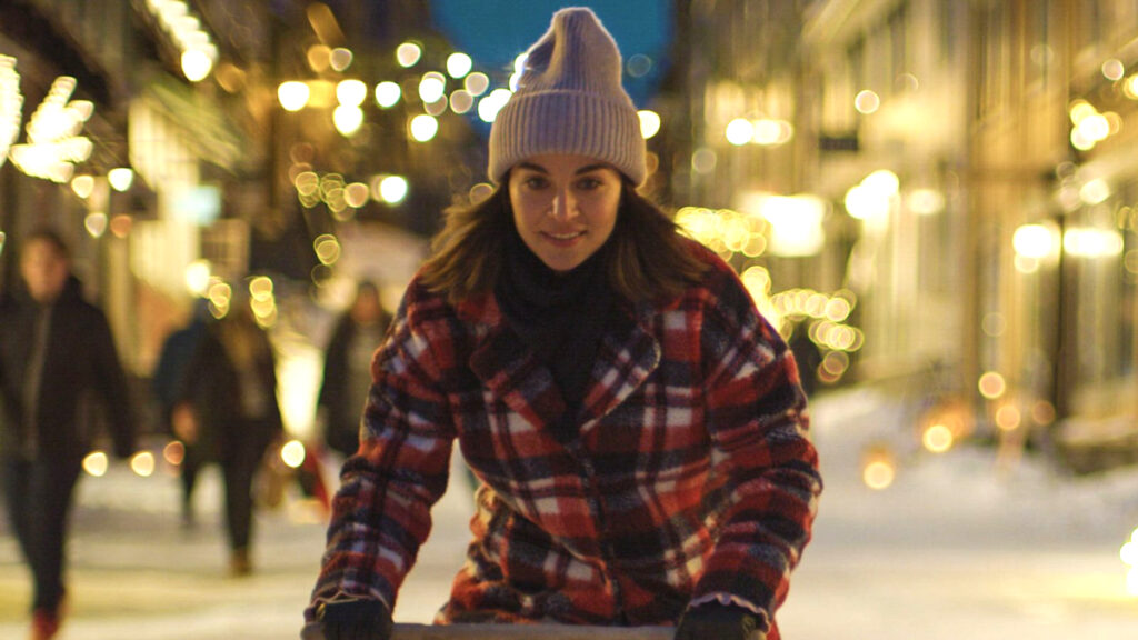 Eine Person in einem rot-schwarz karierten Mantel und Mütze, steht auf einer winterlichen, von Lichtern geschmückten Strasse. Hintergrund unscharf mit weiteren Fussgängern und festlicher Beleuchtung in einer städtischen Umgebung bei Abenddämmerung.