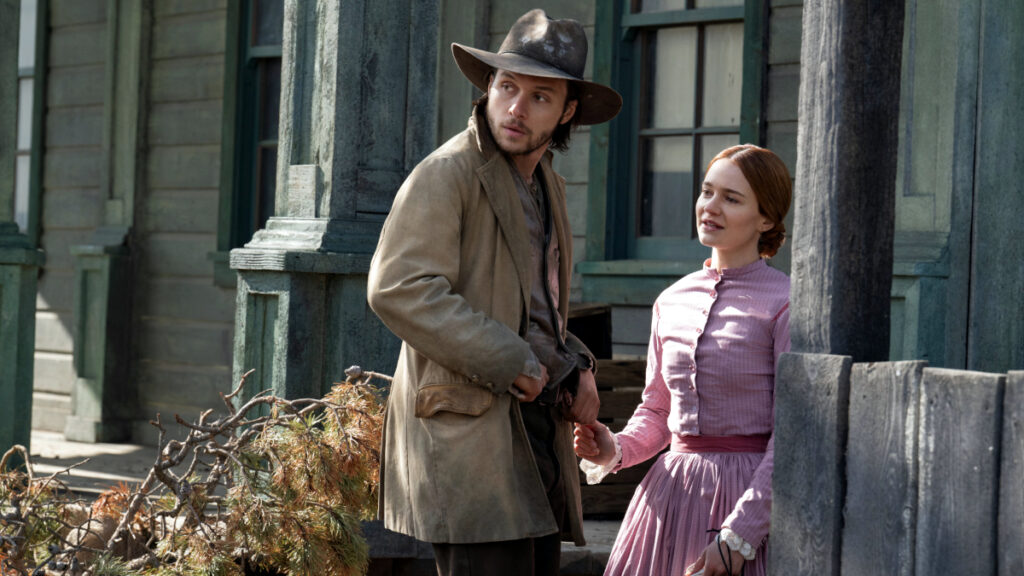Ein Mann mit Hut und eine Frau in einem rosa Kleid stehen vor einem Holzhaus. Der Mann schaut nach links, während die Frau seine Hand hält und ihn anschaut. Beide stehen neben einem Stapel Äste und scheinen in ein Gespräch vertieft zu sein. Die Szene wirkt historisch und ländlich.