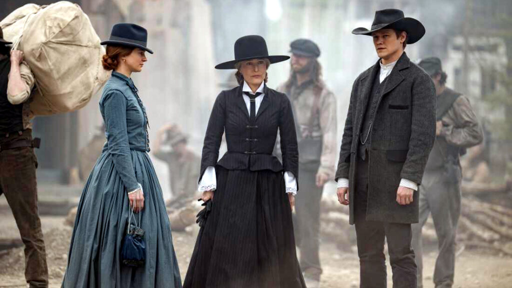 Drei Personen in historischer Kleidung stehen auf einer staubigen Strasse. Zwei Frauen und ein Mann tragen viktorianische Outfits mit Hüten. Im Hintergrund sind unscharfe Figuren und ein Träger mit einem grossen Sack zu sehen. Die Szene wirkt wie aus einem Western-Drama.