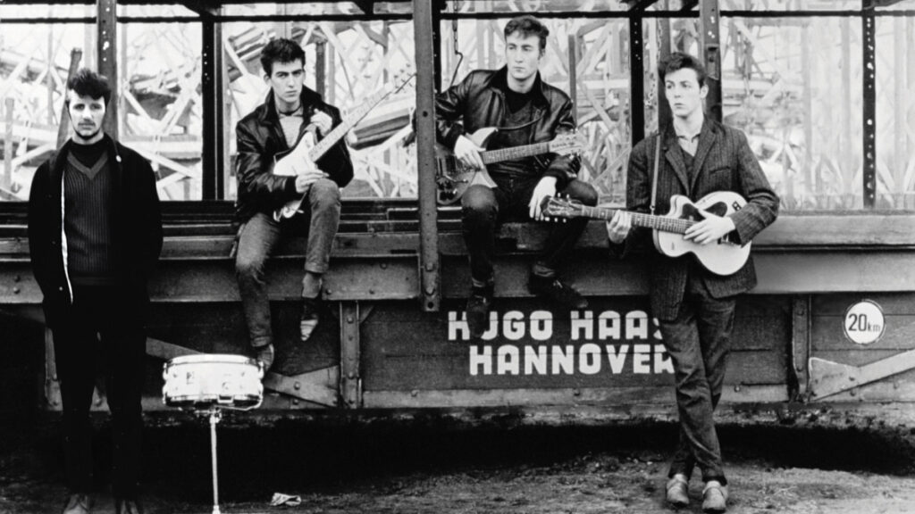 Vier junge Männer posieren in einem schwarz-weissen Foto vor einem alten Güterwagen. Drei halten Gitarren, während einer steht. Eine Trommel steht vor ihnen. Das Bild hat einen Vintage-Look und die Männer tragen Lederjacken und Anzüge.