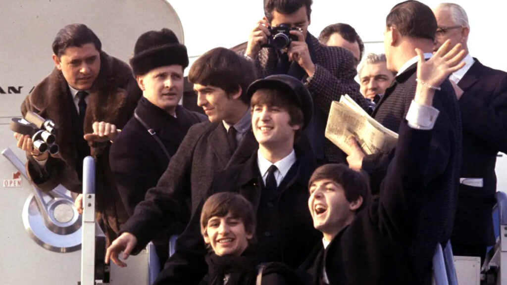 Eine Gruppe junger Männer in Anzügen steht auf einer Flugzeugtreppe. Im Hintergrund sind Personen mit Fotoapparaten und Zeitungen. Alle wirken fröhlich und einige winken. Die Szene vermittelt eine lebhafte und positive Stimmung.