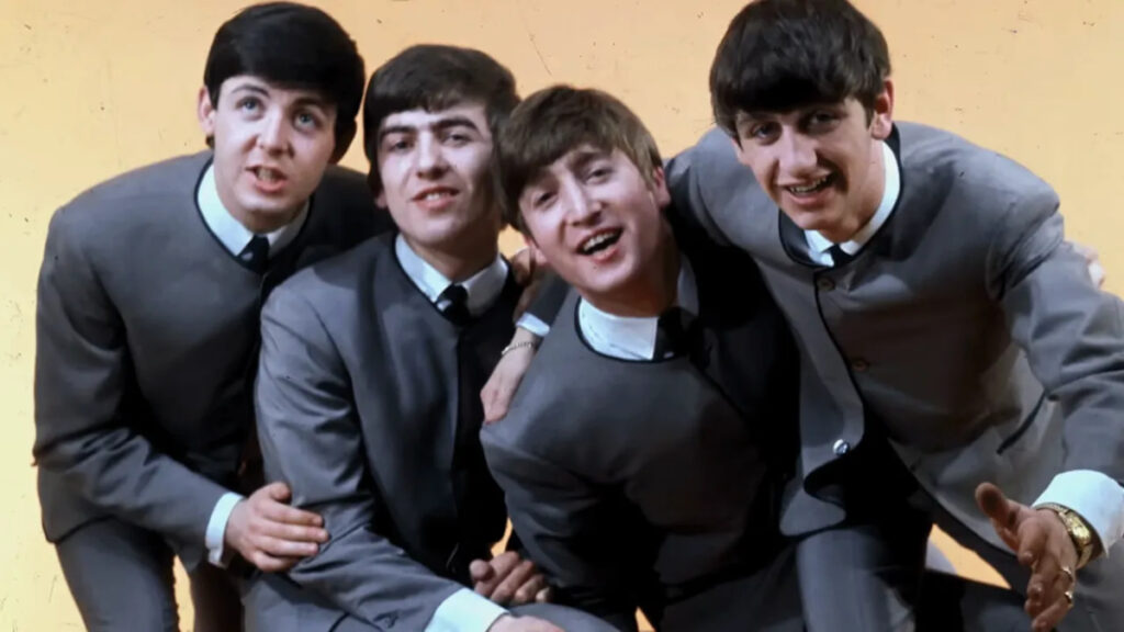 Vier junge Männer in grauen Anzügen posieren fröhlich vor einem gelben Hintergrund. Sie sind nah beieinander, lächeln und haben die Arme umeinander gelegt. Alle tragen weiss-schwarze Hemden und Krawatten. Die Atmosphäre wirkt lebendig und freundschaftlich.