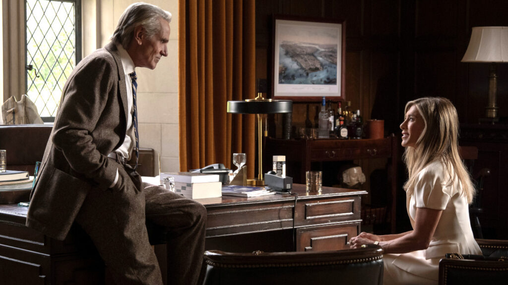 Ein Mann in einem grauen Anzug sitzt auf einem Schreibtisch und spricht mit einer Frau in einem weissen Kleid, die auf einem Stuhl vor ihm sitzt. Der Raum ist mit dunklen Holzmöbeln und einer Tischlampe ausgestattet; im Hintergrund steht eine Bar mit Gläsern und Flaschen.