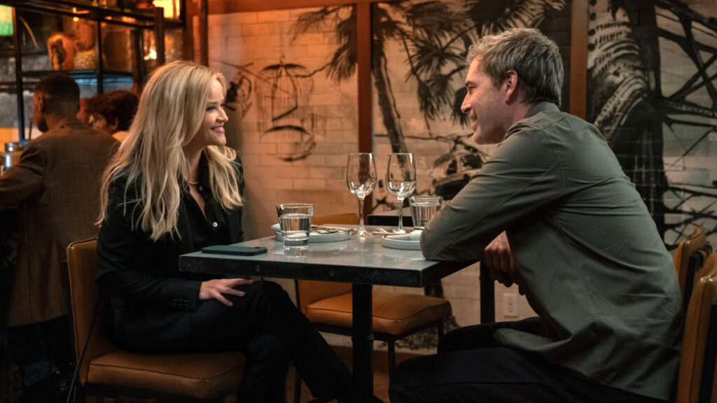 Zwei Personen sitzen sich in einem stilvollen Restaurant an einem Tisch gegenüber und lächeln sich an. Auf dem Tisch stehen leere Weingläser und Wassergläser. Im Hintergrund ist eine Wand mit tropischen Motiven zu sehen. Die Atmosphäre wirkt gemütlich und einladend.