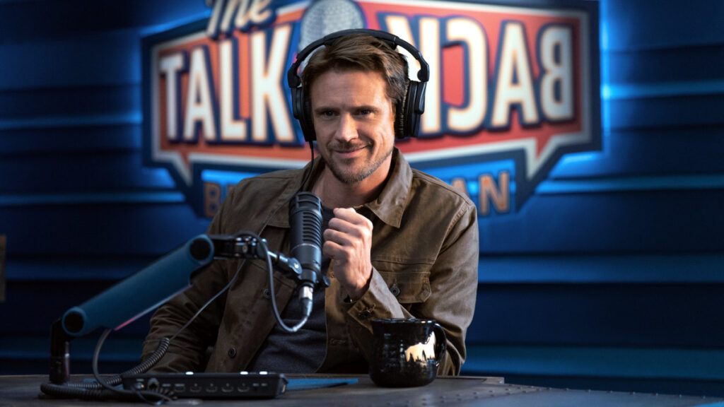 Eine Person sitzt in einem Studio mit Kopfhörern vor einem Mikrofon. Im Hintergrund ist das Schild einer Talkshow zu sehen. Auf dem Tisch steht eine Tasse. Die Atmosphäre wirkt professionell und lässig.