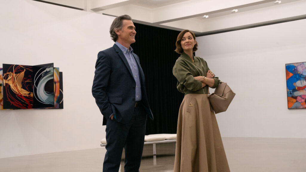 Ein Mann und eine Frau stehen in einer modernen Kunstgalerie. Er trägt einen blauen Anzug, während sie in einem olivgrünen Oberteil und einem langen beigen Rock gekleidet ist. Im Hintergrund sind abstrakte Gemälde an den Wänden zu sehen.