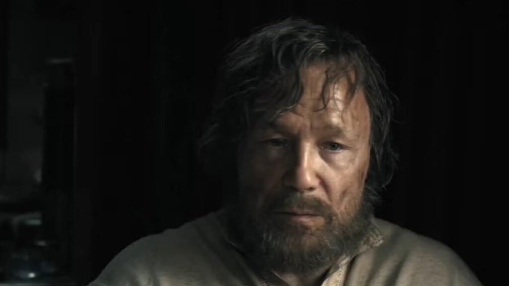 Ein Mann mit ungepflegtem Haar und Vollbart sitzt vor einem dunklen Hintergrund. Sein Gesichtsausdruck wirkt nachdenklich.