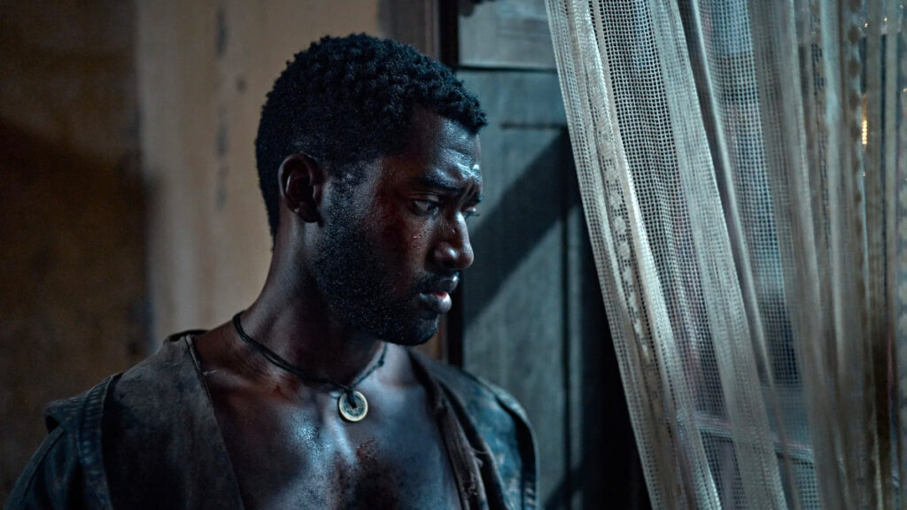 Eine Person mit nachdenklichem Ausdruck steht neben einem Fenster mit transparentem Vorhang. Sie trägt eine einfache Jacke.