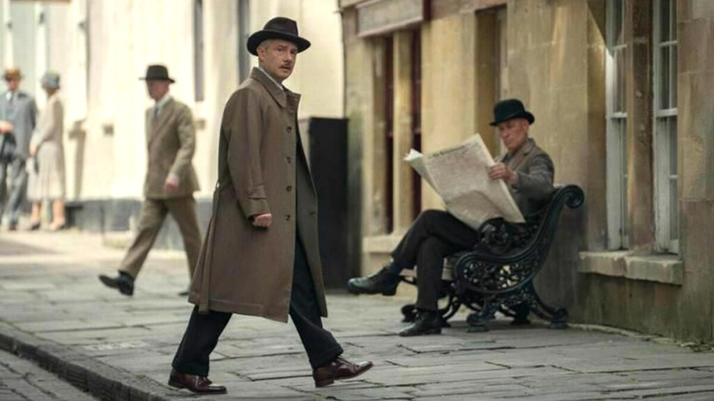 Ein Mann im langen braunen Mantel und Hut geht über eine gepflasterte Strasse in einer historischen Stadtkulisse. Im Hintergrund sitzt eine Person auf einer Bank und liest Zeitung.