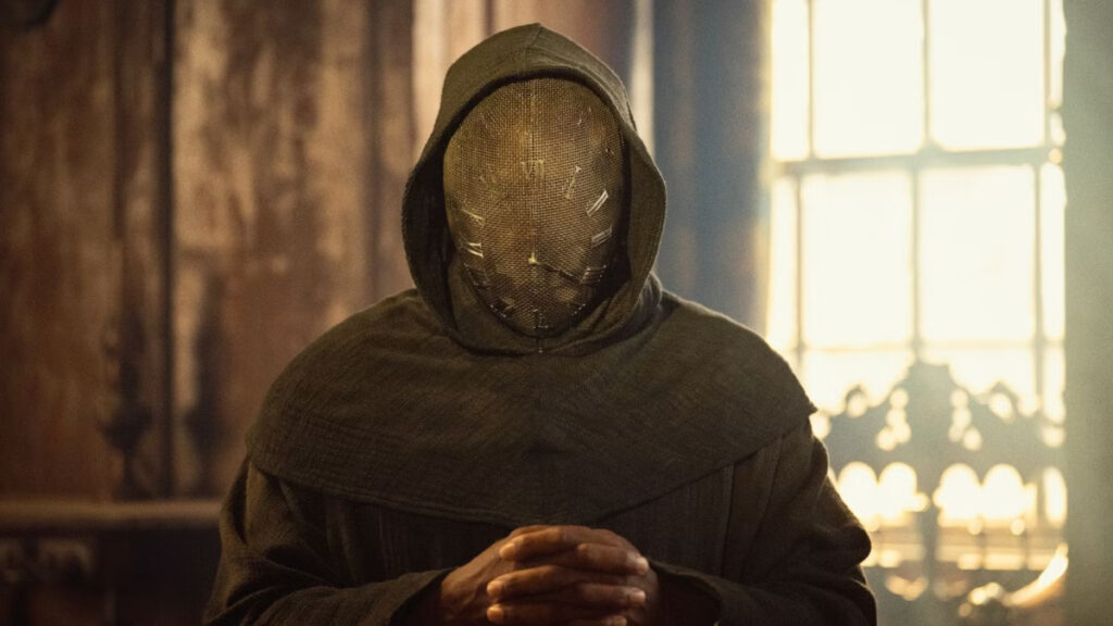 Ein mysteriöser Charakter in dunkler Kapuzenkleidung sitzt in einem warm beleuchteten Raum. Das Gesicht ist vollständig von einer grobmaschigen Maske verdeckt. Im Hintergrund sind verschwommene Fenster und warme Lichtquellen zu sehen, die eine düstere Atmosphäre schaffen.