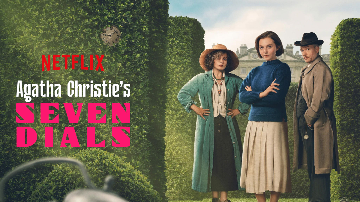 Drei Personen stehen vor einer Kulisse aus Hecken und einem historischen Gebäude. Links erscheint der Schriftzug «Netflix Agatha Christie's Seven Dials». Eine antike Uhr ist im Hintergrund sichtbar. Die Personen tragen historische Kleidung und blicken ernst in die Kamera.