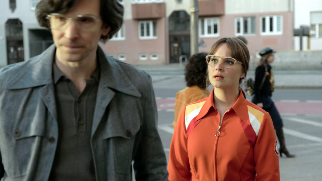 Eine Frau in einer orangefarbenen Jacke und ein Mann in einem grauen Mantel stehen auf einer Strasse in einer städtischen Umgebung. Beide tragen Brillen. Im Hintergrund sind Gebäude und weitere Personen zu sehen.
