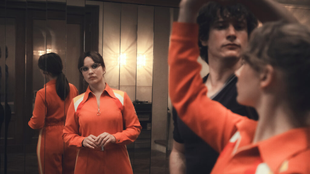 Eine Frau in einem orangefarbenen Trainingsanzug steht vor einem Spiegel. Ihr nachdenklicher Blick und die lockere Haarfrisur dominieren die Szene. Im Vordergrund tanzen unscharf ein Mann und eine weitere Person. Der Raum ist dezent beleuchtet, was eine dramatische Stimmung erzeugt.