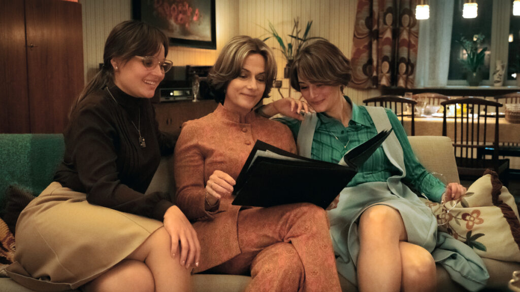 Drei Frauen sitzen auf einem Sofa und betrachten gemeinsam ein offenes Fotoalbum. Die Szene wirkt gemütlich und intim, mit warmem Licht und klassischem Dekor im Hintergrund, darunter Pflanzen und ein gemusterter Vorhang. Alle scheinen zufrieden und vertieft in die Bilder.