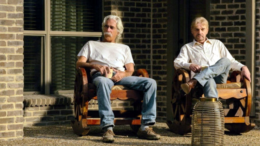 Zwei ältere Männer mit grauen Haaren sitzen entspannt in Holzstühlen vor einem Backsteinhaus. Der linke trägt ein weisses T-Shirt und Jeans, der rechte ein helles Hemd. Zwischen ihnen steht ein kleiner Korb. Die Szene wirkt ruhig und alltäglich.