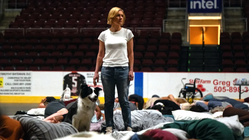 Eine Frau in weissem T-Shirt und Jeans steht in einer Arena. Neben ihr sitzt ein Hund. Um sie herum liegen mehrere Menschen auf Matten oder Decken. Die Tribünen im Hintergrund sind leer.