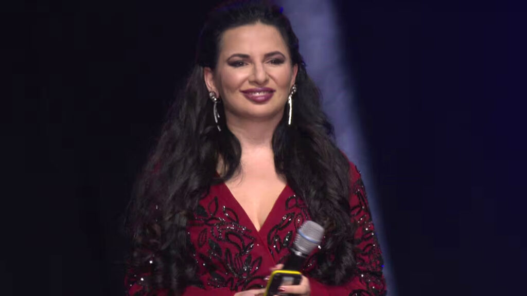Eine Frau mit langen dunklen Haaren steht lächelnd vor einer bühnenartigen Kulisse mit violetter Beleuchtung. Sie trägt ein rotes Kleid mit schwarzen Pailletten-Ornamenten und lange Ohrringe. In der Hand hält sie ein Mikrofon.