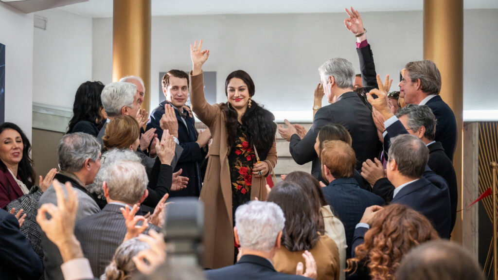 Ein Mann und eine Frau stehen in der Mitte einer applaudierenden Menschenmenge in einem modernen Raum mit goldenen Säulen. Beide winken lächelnd mit erhobenen Händen. Die Atmosphäre wirkt festlich.