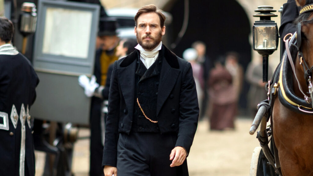 Mann in schwarzem Mantel und weisser Krawatte im historischen Stil steht vor einer historischen Kutsche. 