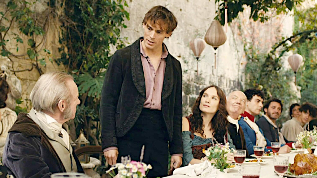 Ein junger Mann mit braunen lockigen Haaren steht in dunklem Anzug an einem gedeckten Tisch im Freien. Links sitzt ein älterer Mann, rechts von ihm eine junge Frau.