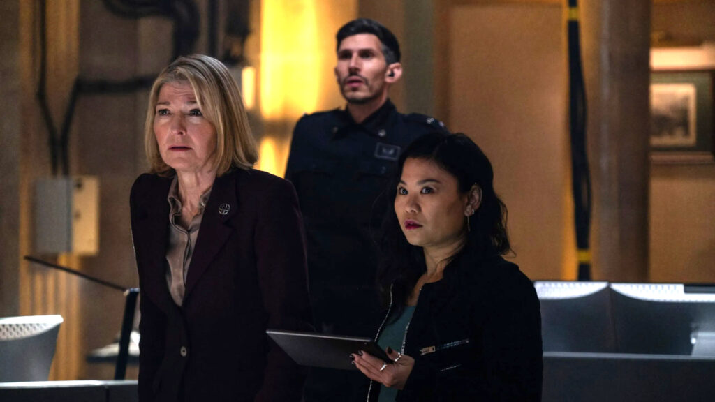 Drei Personen in einem futuristischen Raum wirken angespannt. Eine Frau mittleren Alters trägt einen dunkelroten Blazer, eine jüngere Frau hält ein Tablet, und ein Mann in Uniform steht im Hintergrund. Die Beleuchtung ist warm und die Umgebung wirkt technisch-modern.