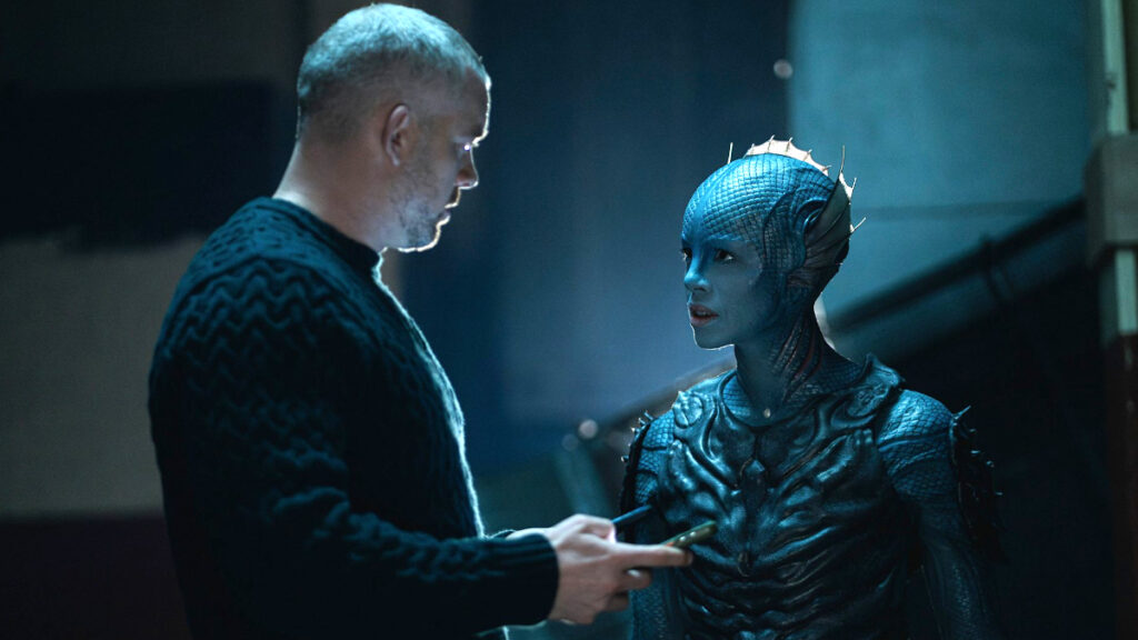 Ein Mann in einem dunklen Pullover spricht mit einer blauhäutigen, ausserirdisch wirkenden Figur in aufwendigem Kostüm. Beide stehen in einer gedämpft beleuchteten Umgebung, während der Mann ein Gerät in der Hand hält, auf das er zeigt.