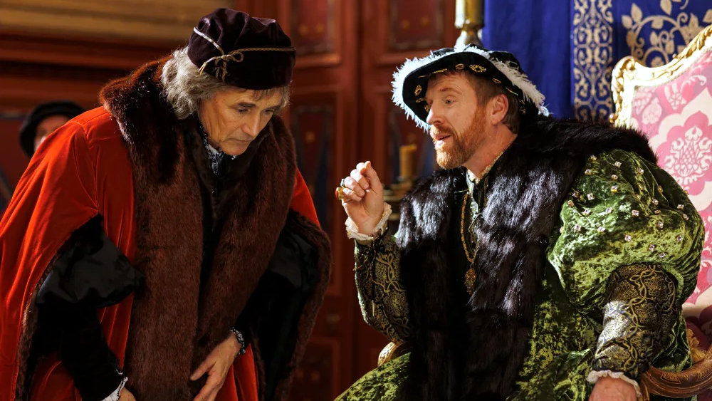 Zwei Männer in historischer Kleidung. Der linke trägt einen roten Mantel und einen Hut, beugt sich leicht vor. 