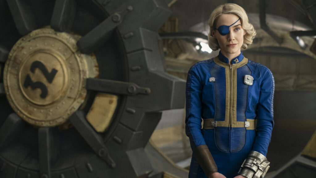 Eine Person mit kurzen blonden Haaren und blauer Augenklappe trägt einen blauen Uniformoverall mit goldenen Akzenten in einem futuristischen Innenraum. Im Hintergrund sind metallische Strukturen und technische Ausrüstung zu sehen.