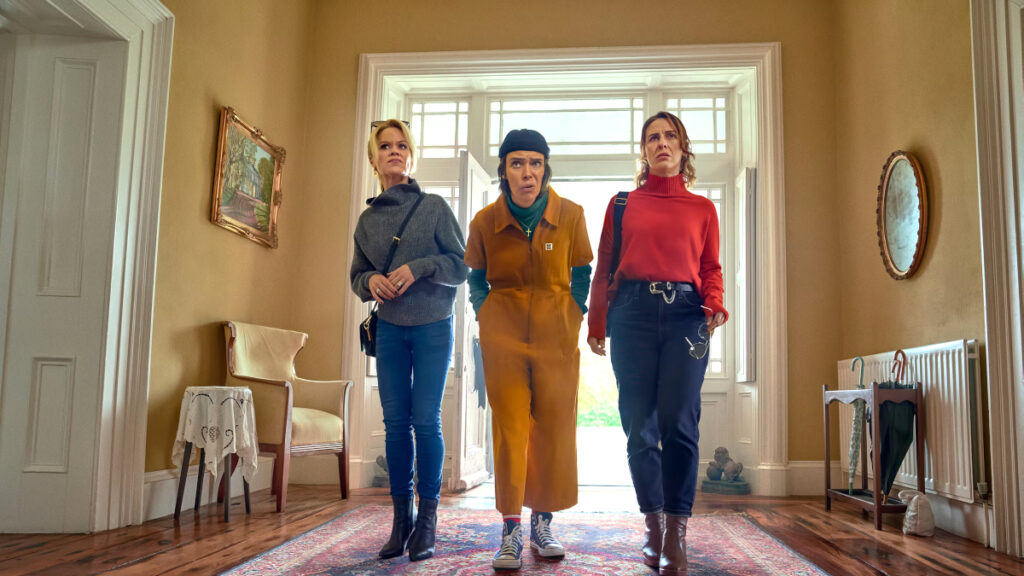 Drei Frauen stehen nebeneinander in einem elegant eingerichteten Wohnzimmer mit Parkettboden und orientalischem Teppich. Von links: Frau in grauem Pullover und Jeans, Frau in orangem Overall mit blauer Mütze, Frau in rotem Pullover und dunkler Hose. Alle blicken ernst in die Kamera.
