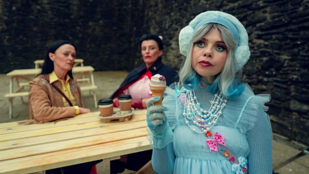 Eine Frau in hellblauem viktorianischem Kleid mit Perlenketten und Häubchen hält eine Eistüte und blickt ernst in die Kamera. Im Hintergrund sitzen zwei weitere Personen an einem Holztisch mit Kaffeetassen in einem Café oder Restaurant.