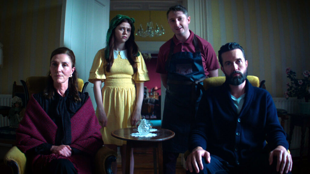Vier Personen in einem Wohnzimmer: Zwei Frauen sitzen links (eine in rotem Mantel, eine in gelbem Kleid mit grünem Kopftuch), zwei Männer stehen rechts (einer in rotem Hemd mit Schürze, einer in blauem Cardigan). Gespannte Atmosphäre, gedämpfte Beleuchtung.