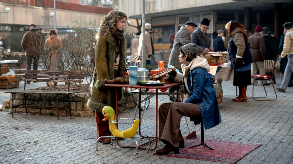 Eine Frau in grünem Mantel steht neben einem kleinen roten Tisch, an dem eine andere Frau in blauem Mantel sitzt. Die Szene spielt auf einem Marktplatz mit Kopfsteinpflaster, im Hintergrund sind weitere Menschen und Marktstände zu sehen. Unter dem Tisch liegt ein gelbes Spielzeug.