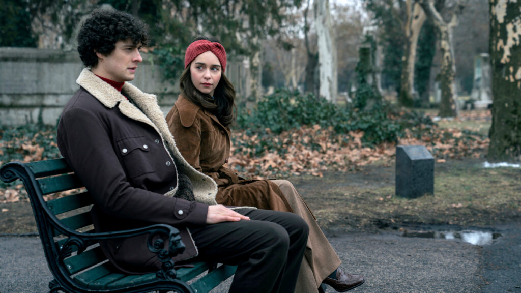 Ein junger Mann mit dunklen Locken in brauner Jacke mit Fellkragen sitzt neben einer Frau mit roter Strickmütze und braunem Mantel auf einer Parkbank. Die herbstliche Szene zeigt Bäume und herabgefallene Blätter im Hintergrund.