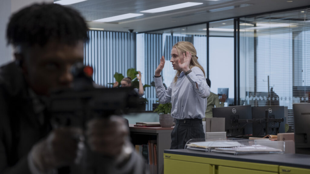 Eine blonde Frau in weisser Bluse und dunklem Rock steht mit erhobenen Händen in einem modernen Grossraumbüro. Im Vordergrund ist eine Person unscharf zu sehen, die eine Waffe im Anschlag hält.