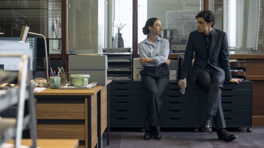 Zwei Personen in einem modernen Büro: Eine Frau in hellblauem Hemd und ein Mann in dunklem Anzug sitzen nebeneinander auf einem schwarzen Schreibtisch. Im Hintergrund sind Fenster mit Jalousien, Büromöbel und ein Computer zu sehen.