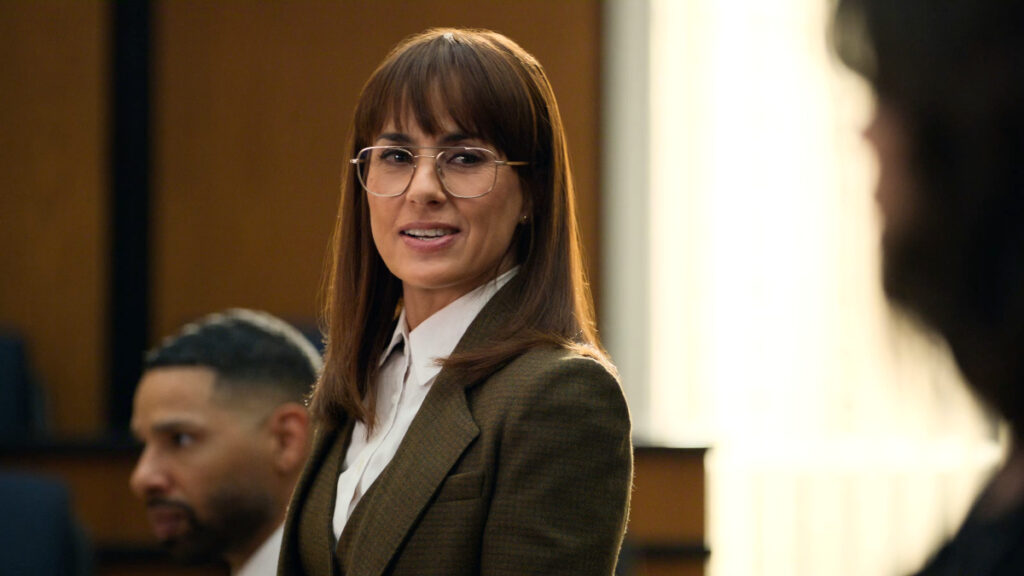 Eine Frau mit braunen Haaren und Brille trägt einen olivgrünen Blazer über einem weissen Hemd. Sie steht in einem Gerichtssaal und lächelt leicht. Im Hintergrund sind unscharf weitere Personen zu sehen.