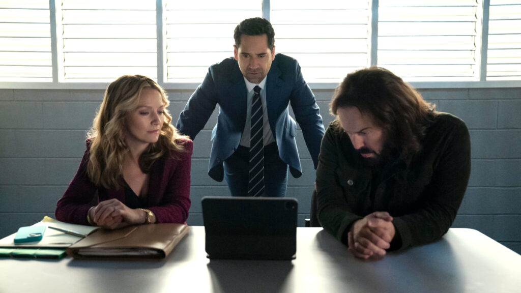 Drei Personen in einem Verhörraum oder Büro: Eine blonde Frau in burgunderrotem Blazer und ein Mann mit längeren Haaren sitzen an einem Tisch mit Laptop und Akten, während ein Mann im dunklen Anzug hinter ihnen steht. Jalousien im Hintergrund lassen Licht herein.