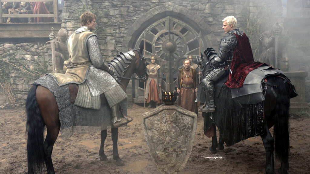 Mittelalterliche Szene mit zwei Reitern in Rüstung auf Pferden, die sich in einem Burghof gegenüberstehen. Im Hintergrund sind weitere Personen vor einer Steinmauer mit rundem Fenster zu sehen. Die Atmosphäre wirkt dramatisch und spannungsgeladen.