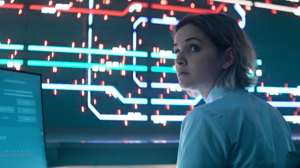 Eine blonde Frau in weisser Kleidung steht vor einer futuristischen Kulisse mit leuchtenden blauen und roten Neonlinien. Die Szenerie zeigt komplexe elektronische Schaltkreismuster im Hintergrund.