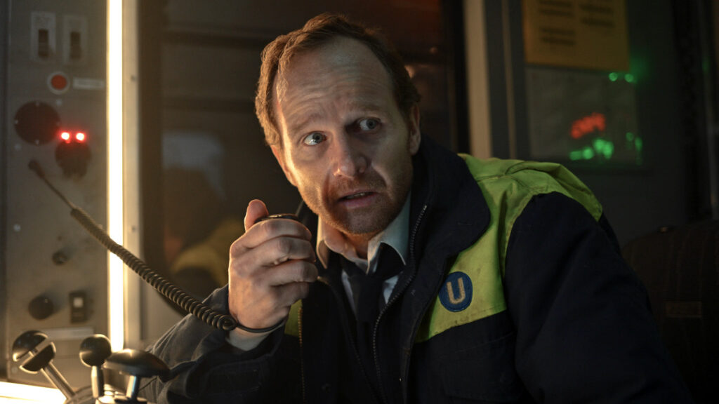 Ein Mann mit Bart in einer schwarzen Jacke mit grünen Reflektorstreifen spricht in ein Handfunkgerät. Er befindet sich im Führerstand einer U-Bahn mit Kontrollpanels und roten sowie grünen Statusleuchten im Hintergrund.