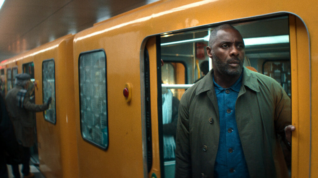 Ein Mann in olivgrüner Jacke und blauem Hemd steht vor einem gelben U-Bahn-Zug in einer unterirdischen Station. Er blickt ernst zur Seite, während sich die Türen des beleuchteten Waggons öffnen. Die Szene wirkt cinematographisch und spannungsgeladen.